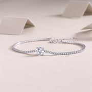 A silver bracelet featuring a line of round-cut moissanite stones with a central round-cut moissanite stone, placed on a beige surface.